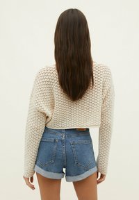 Mujer con cabello largo y castaño, vistiendo un suéter de punto beige y pantalones cortos de mezclilla azul con puños, de pie contra un fondo claro y liso.