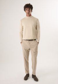 Beige gebreide coltrui met geribbelde schouderdetails, gecombineerd met lichtbeige broek en bruine suède enkellaarsjes. Eenvoudig ontwerp.