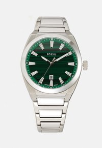 Montre Fossil avec un cadran rayé vert, des index en argent, une fonction date à 6 heures et un bracelet en acier inoxydable avec des accents blancs.