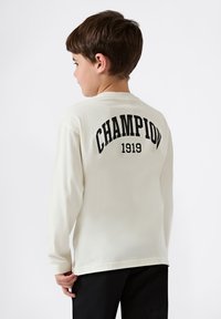 Kremowa koszula z długim rękawem z czarnym napisem "CHAMPION" oraz "1919" poniżej z tyłu, o luźnym kroju i okrągłym dekolcie.
