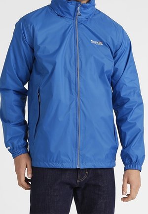 Blaue Outdoor-Jacke mit Reißverschluss, elastischen Bündchen, zwei seitlichen Reißverschlusstaschen und kleinem Logo auf der Brust, getragen über dunklen Jeans.