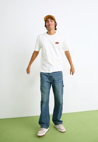 Hvit t-skjorte med kort erme og rødt grafisk trykk, blå vide jeans, beige sneakers, og en lys brun caps, på en ensfarget bakgrunn.