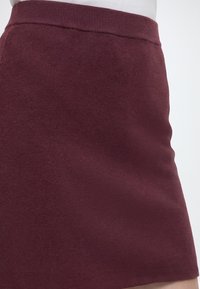 Gros plan d'une personne portant une jupe en maille unie bordeaux avec une taille côtelée, associée à un haut blanc.