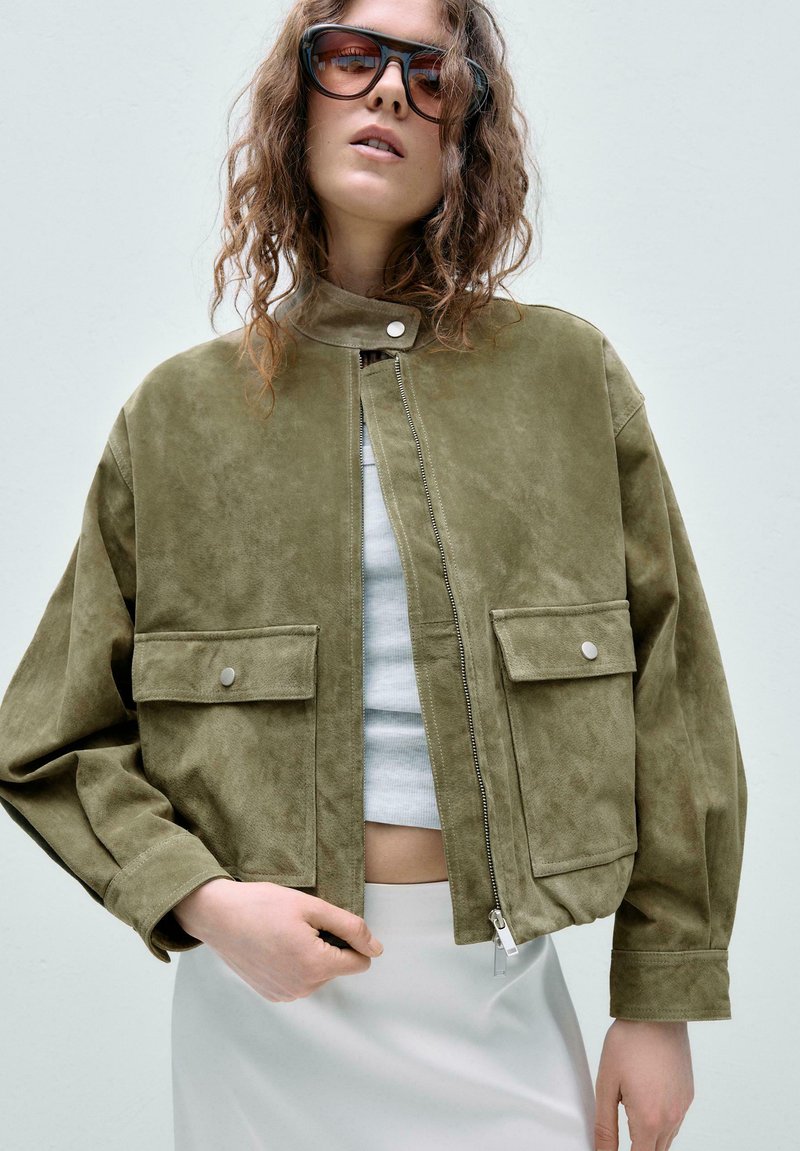 Femme portant une veste zippée en daim vert avec poches à rabat, un crop top blanc, une jupe blanche et des lunettes de soleil oversize devant un fond uni.