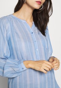 Blusa manica lunga blu chiaro con righe bianche verticali, patta con bottoni e taschino sul petto. Tessuto morbido e texturizzato.