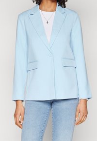Světle modrý blazer s přiléhavým střihem, zapínání na jeden knoflík, klopové límce a dvě přední kapsy. Nosený přes bílý top a džíny.