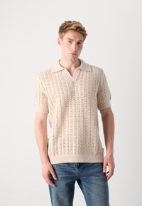 Knit polo shirt in light beige with a textured pattern, short sleeves, and a soft collar. Pairing with blue denim jeans.