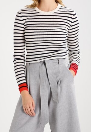 Nainen pukeutunut mustavalkoraidalliseen pitkähihaiseen yläosaan, jossa on punaiset kalvosimet, yhdistettynä korkeavyötäröisiin vaaleanharmaisiin leveälahkeisiin housuihin ja solmittuun vyöhön.