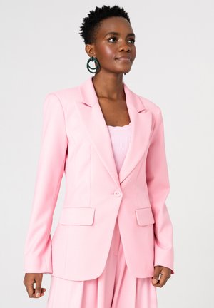 Kobieta o krótkich kręconych włosach, nosząca duże ciemnozielone kolczyki-koła i jasnoróżową marynarkę na prążkowanym topie, stojąca na jednolitym tle.