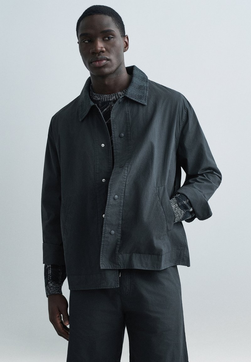 Czarna lekka kurtka z ostrym kołnierzem, zatrzaskami i bocznymi kieszeniami. Noszona na teksturowanym ciemnym swetrze i dopasowanych spodniach.