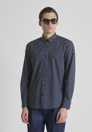 Man draagt zwarte zonnebril en een donkerblauw overhemd met witte streepjes, staand tegen een effen lichte achtergrond.