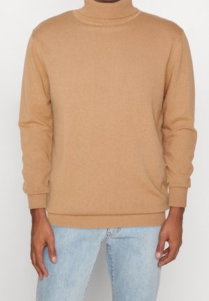 Pull col roulé beige à texture côtelée et manches longues, avec un col et des poignets ajustés. Porté avec un jean bleu clair.