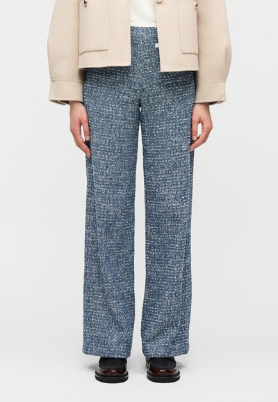 Γυναίκα που φοράει μπλε υφασμάτινη παντελόνα με φαρδιά πόδια, μπεζ σακάκι, λευκό τοπ και σκούρα καφέ παπούτσια, στέκεται μπροστά σε απλό φόντο.