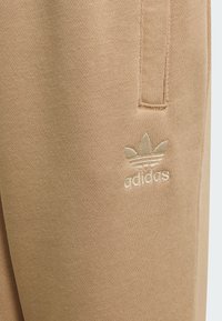 Béžové teplákové nohavice z jemnej tkaniny s vyšívaným logom Adidas v svetlejšom odtieni, rovného strihu a s detailom bočného vrecka.