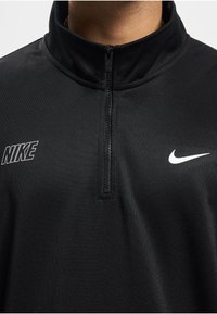 Čierna športová mikina s štvrťovým zipsom, s logom Nike na pravej strane hrude a bielym znakom Nike na ľavej strane hrude.