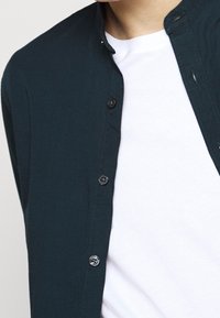 Ciemnoniebieska koszula z guzikiem i stójką, wykonana z teksturowanej tkaniny i czarnymi guzikami. Nosi się ją na gładkiej białej koszulce.