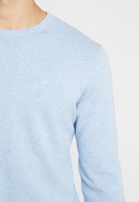 Light blue, long-sleeve sweater in a soft knit with a round neck. Subtle logo detail on the chest. Smooth texture, fitted design.