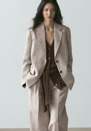 Frau in einem beige übergroßen Anzug mit braunem, geknöpftem Strickweste und Bindegürtel, stehend mit den Händen in den Taschen vor einfachem Hintergrund.