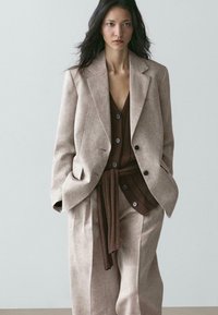 Žena v béžovém oversize obleku s hnědou pletenou vestou na knoflíky a opaskem, stojící s rukama v kapsách na jednobarevném pozadí.