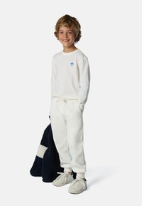 Felpa bianca con logo blu, pantaloni jogger bianchi e sneakers beige, in mano una giacca in pile blu scuro con un motivo chiaro.