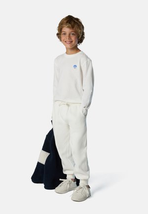 Felpa bianca con logo blu, pantaloni jogger bianchi e sneakers beige, in mano una giacca in pile blu scuro con un motivo chiaro.