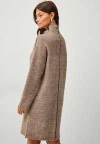 Gebreide jurk in beige met een hoge hals, voorzien van een zwarte verticale streep langs de achterkant, lange mouwen en een relaxte pasvorm.