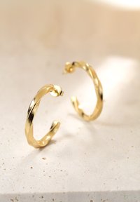 Pendientes de aro de oro con un diseño retorcido, que cuentan con una superficie pulida y un cierre seguro. Sobre un fondo texturizado neutro.