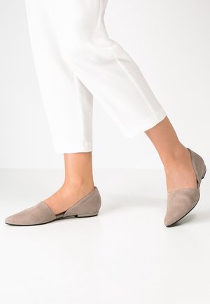 Person trägt beige Wildleder-Spitzenschuhe mit niedrigen Absätzen und weiße gekürzte Hosen, steht auf einem glatten weißen Boden.