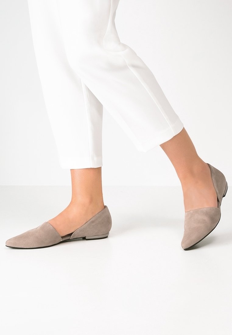 Personne portant des ballerines en daim beige à bout pointu avec des talons bas et un pantalon blanc coupé, debout sur un sol blanc lisse.