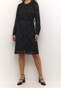 Svart spetsklänning med långa ärmar, knäppning framtill och midjeband. Har intrikata blommönster och en scalloped fåll.