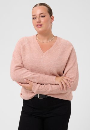Femme à la peau claire et aux cheveux blonds, vêtue d’un pull rose et d’un pantalon noir, debout avec les bras croisés devant un fond clair.
