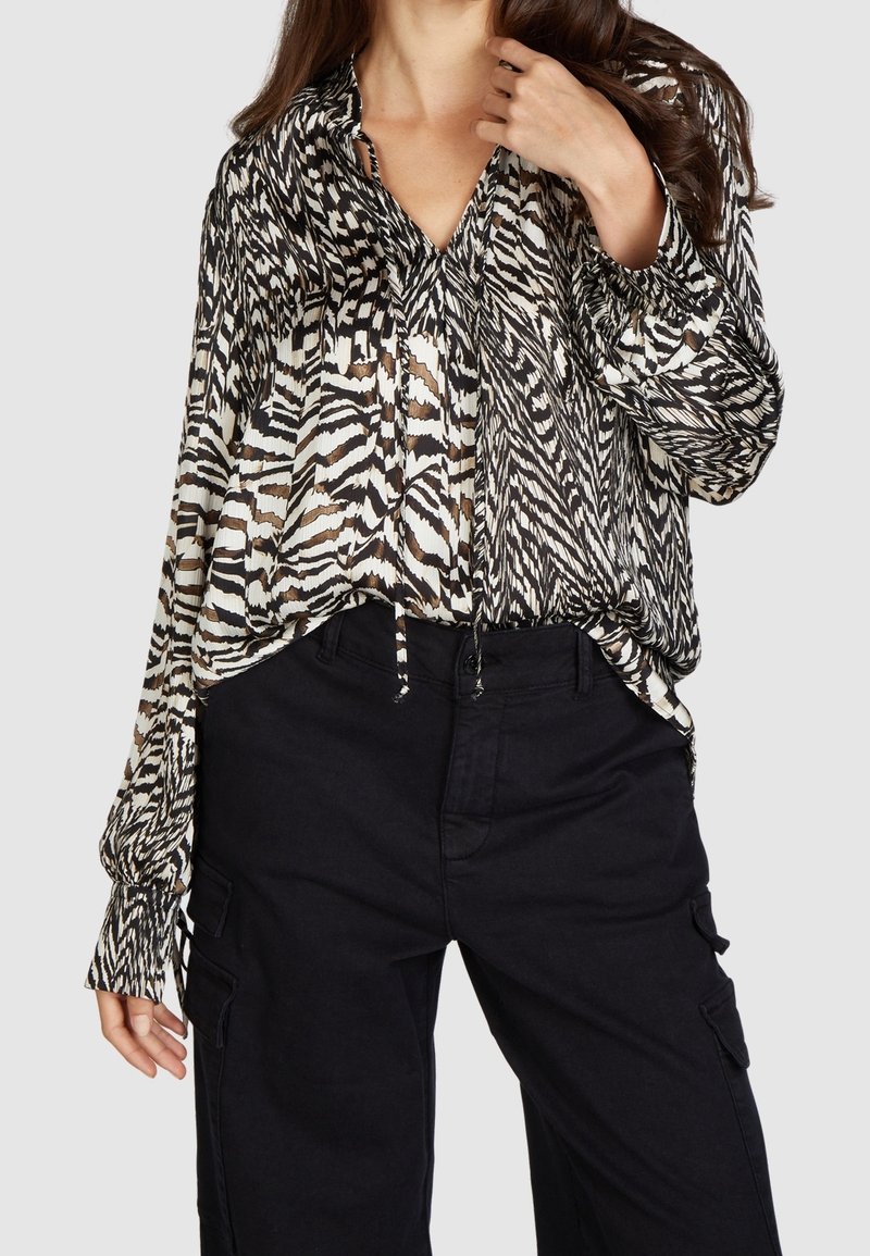 Femme portant un chemisier à manches longues noir et blanc à motifs et un pantalon noir taille haute, avec une main près du cou.