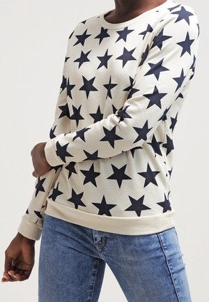 Suéter de manga larga crema con grandes estrellas marinas, cuello redondo y puños acanalados, combinado con jeans de color azul claro. Tejido suave.