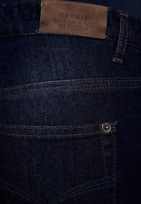 Tmavě modré džíny z denimu s texturovaným povrchem. Mají hnědou koženou etiketu, zlaté prošití a kovové tlačítko na kapse.