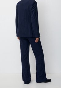 Personne portant un costume bleu marine à fines rayures avec un pantalon à jambes larges et des chaussures noires, debout devant un fond blanc uni.