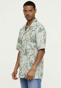 Chemise à manches courtes avec des boutons, de couleur vert clair, avec un motif floral en blanc, jaune et bleu. Tissu texturé avec une coupe décontractée.