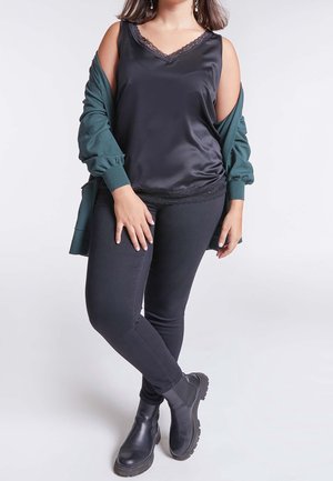 Top en satin noir sans manches avec finition en dentelle, superposé sous un cardigan vert foncé. Associé à un jean noir ajusté et des bottines noires.