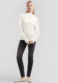 Biały prążkowany sweter z długim rękawem i wysokim kołnierzem, połączony z ciemnoszarymi dopasowanymi jeansami i jasnymi płaskimi butami z wycięciami.