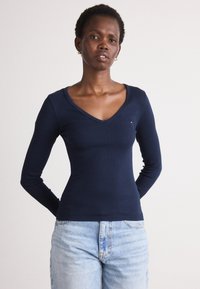 Cămășă lungă cu mâneci, de culoare bleumarin, cu design în V, textură ribbed și detalii cu un logo mic pe piept. Asortată cu blugi din denim deschis.