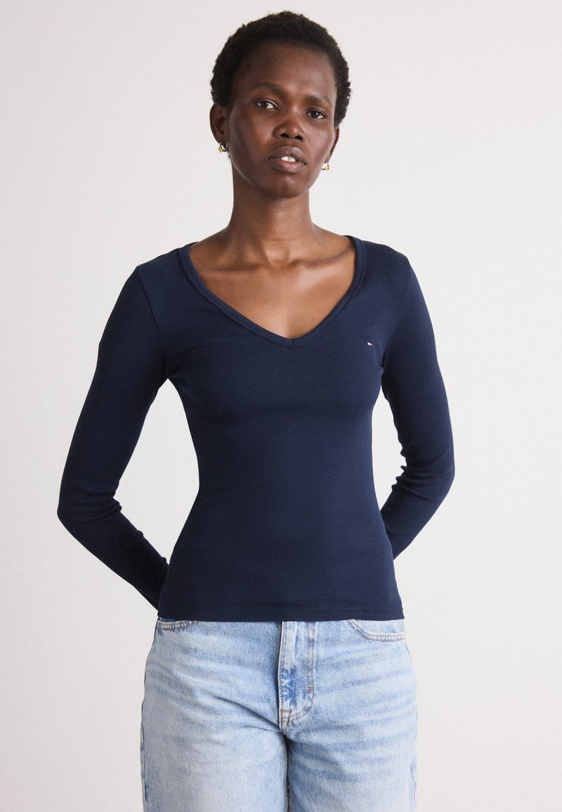 Cămășă lungă cu mâneci, de culoare bleumarin, cu design în V, textură ribbed și detalii cu un logo mic pe piept. Asortată cu blugi din denim deschis.