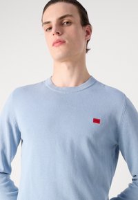 Helles blauer Rundhalsausschnitt-Pullover aus weichem Stoff mit langen Ärmeln und einem kleinen roten Logo-Patch auf der linken Brust.