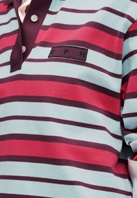 Polo rayé rouge, bleu clair et bordeaux foncé avec deux boutons et un patch rectangulaire marqué "CPH" sur la poitrine.