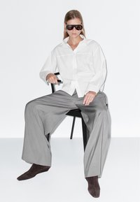 Camicia bianca abbottonata, pantaloni grigi a gamba larga, stivaletti marroni e occhiali da sole. Modella seduta su una sedia nera su sfondo bianco.