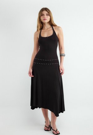 Młoda kobieta ubrana w czarną sukienkę z wiązaniem na szyi z srebrnymi ćwiekami, asymetrycznym dołem, czarne paski na obcasie i srebrną bransoletką na ramieniu.