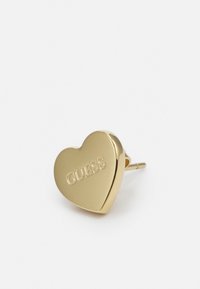 Orecchino a forma di cuore in oro con logo "GUESS" inciso, superficie metallica liscia e chiusura a perno per una fissatura sicura.