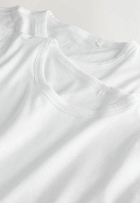 Divas baltas kokvilnas T-krekli sakrautas, ar apaļu kakla izgriezumu, īsām piedurknēm un plakanām šuvēm ar gludu, mīkstu tekstūru.