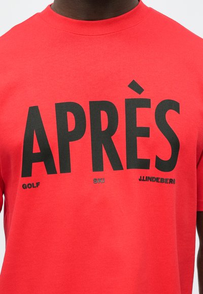 Κόκκινη μπλούζα με μεγάλο μαύρο κείμενο "APRÈS" και μικρότερες λέξεις "GOLF", "SKI" και "J.LINDEBERG" τυπωμένες κάτω από το στήθος.