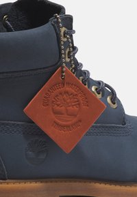 Stivale impermeabile navy con tomaia in pelle, occhielli in metallo e lacci in tessuto. Presenta un’etichetta arancione testurizzata che mostra il logo Timberland.