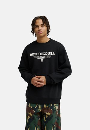 Person steht, trägt ein schwarzes DC Shoes Sweatshirt mit weißem Schriftzug und grüne Camouflage-Hose, Hände gefaltet, neutraler Gesichtsausdruck.