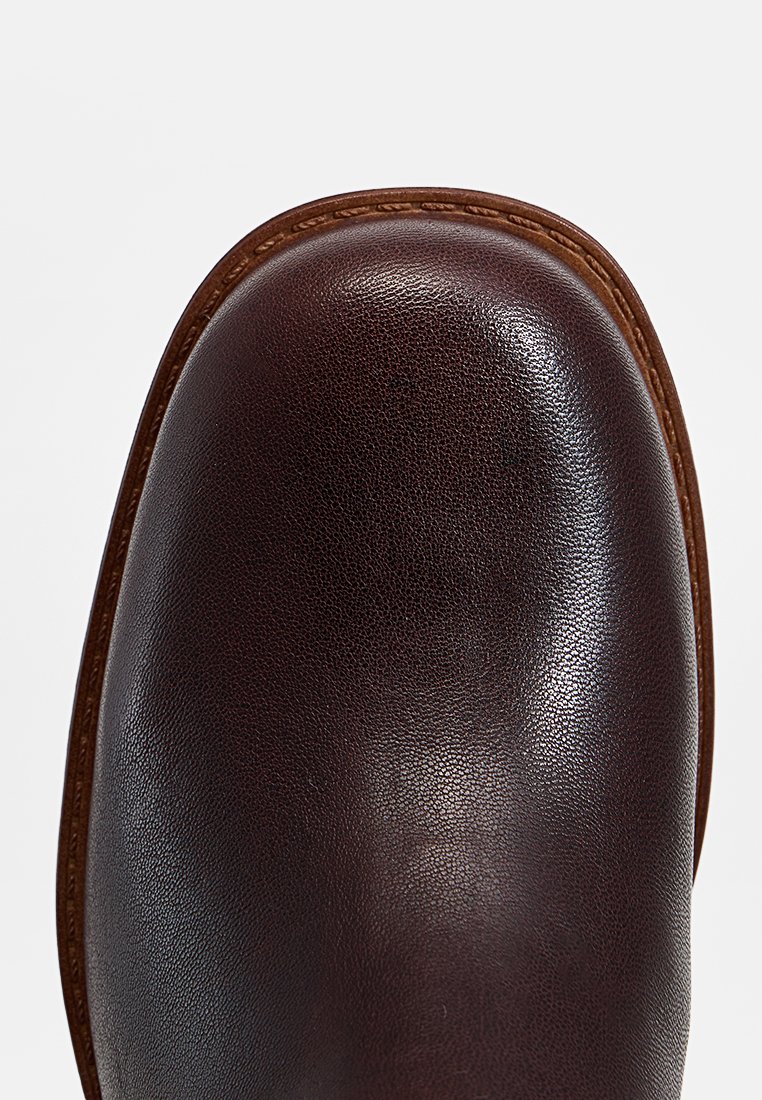 Bota de cuero marrón con textura suave, punta redondeada y costuras en contraste. Presenta una suela resistente y un acabado pulido.